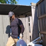 A man wearing a black shirt, tan pants, gloves, and a cap carries a large bag of trash outside near a wooden fence with piles of debris and boards behind him on a sunny TX day, highlighting trash removal in Dallas.