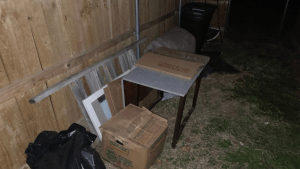 A wooden fence at night with cardboard boxes, a small table, panels, a black trash bag, and other miscellaneous items stacked on the grass—perfect for junk removal in Arlington, TX by Arlington Junk Removal Service.