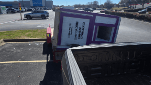 A large clothing donation box tipped over onto its side in a Dallas, TX parking lot near a pickup truck, with shopping centers and parked cars in the background on a sunny day—perfect for donation box removal in Dallas, TX.
