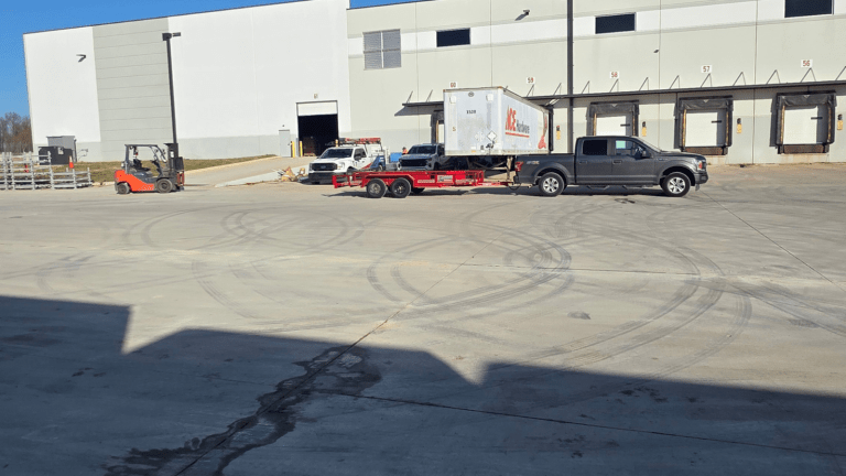 A forklift, a pickup truck with a trailer for junk removal, and two utility vehicles are parked outside a large warehouse with multiple loading docks on a sunny day. Tire marks are visible on the concrete surface.