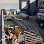 A loading dock area with metal pipes, wooden pallets, and scrap metal ready for removal in Wilmer, TX by Arlington Junk Removal Service. Cardboard boxes and various construction materials are scattered nearby; a van is parked at numbered dock doors 59 and 60.