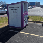 A purple-and-white clothing and shoe recycling bin, ideal for donation box removal in Dallas, sits in a parking lot near Sam’s Club. Cars and a trailer are parked nearby under a clear, sunny sky.