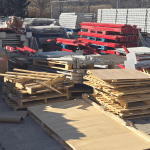 Stacks of wooden pallets and various metal construction materials, including red beams and metal sheets, await scrap metal removal in Dallas at an organized outdoor storage area beside a chain-link fence on a sunny day.