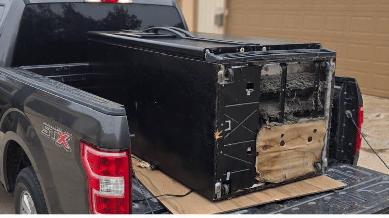 A large, old, black appliance, possibly a refrigerator, lies on its back in the bed of a dark-colored pickup truck—likely ready for refrigerator removal in Euless by Arlington Junk Removal Service. The worn unit has exposed wiring and insulation on its bottom panel.