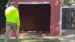 A person in a bright yellow shirt and khaki pants walks toward a small red shed with peeling white trim, ready for shed removal by Arlington Junk Removal Service. The empty shed’s open doorway reveals debris scattered on the ground.