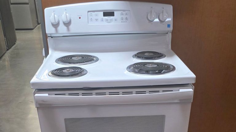 A white electric stove with four coil burners and an oven, perfect for Appliance Removal & Disposal in Arlington, is positioned against a wooden wall and reflective floor, featuring control knobs and a digital display panel on the back.