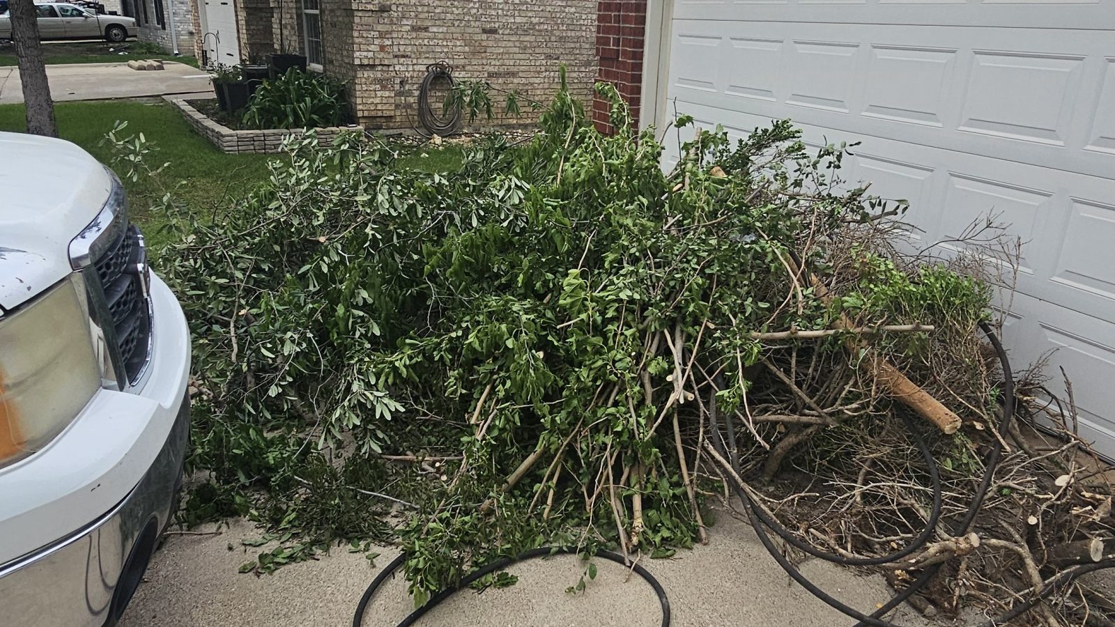 A large pile of tree branches and yard debris is stacked on a driveway in front of a garage at a suburban house, awaiting yard debris removal by Arlington Junk Removal Service. A white vehicle is parked nearby, and a garden hose lies on the ground.